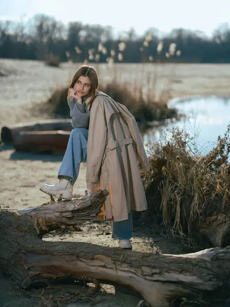 Trençkotlu genç kadın sahilde duruyor odunların ve gölün kıyısında sakin suların yansıttığı gökyüzü ve baharın başlarındaki bulutları..