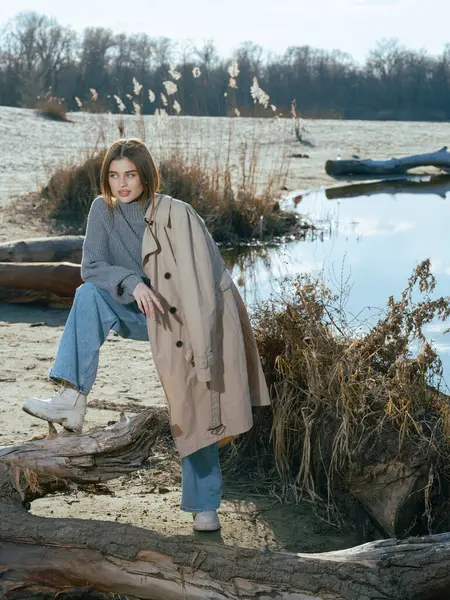 Trençkotlu genç bir kadın kumsalda, odunların ve gölün kenarında duruyor. Sakin suları gökyüzünü, bulutları ve baharın başında çıplak ağaçlarla çevrili..