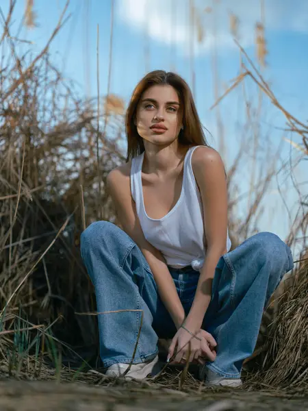 Beyaz kolsuz tişörtlü genç bir kadın ve kuru kamışların arasında oturmuş mavi gökyüzü ve bulutların arkasında gevşeyen bir kot pantolon. Yakın plan. Alt görünüm.