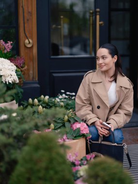 Ceketli ve kotlu gülümseyen genç bir kadın çiçekçinin yanında oturmuş sepetlerdeki çiçek açan ortancalara hayran..