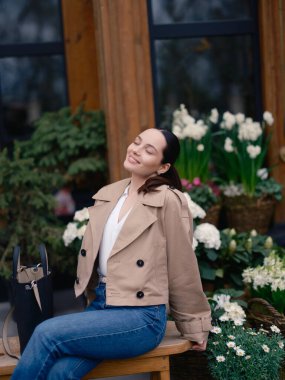 Ceketli ve kot pantolonlu gülümseyen genç bir kadın, bankta oturuyor, dinleniyor ve kapalı gözlerle sepetlerdeki çiçekleri açarak eğleniyor. Yakın plan..