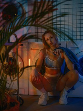 İç çamaşırlı genç sarışın kadın modern çamaşır makinesinin yanında oturuyor ve dinleniyor fayanslı duvarları ve neon ışıkları var. Moda ve günlük hayatın karışımı şık bir atmosfer..