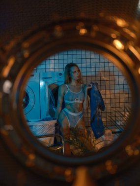 Çamaşır makinesinin kapısından çamaşır makinesinin yanında duran genç sarışın kadına ve modern çamaşırlarda yıkanacak çamaşırları sıralayan kadına bakın. Çamaşırları çamaşır makinesine koyuyorum..