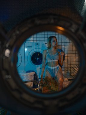 Çamaşır makinesinin kapısından çamaşır makinesinin yanında duran ve çamaşırları modern çamaşırlarda yıkamak için sıralayan genç sarışın kadına bakın..