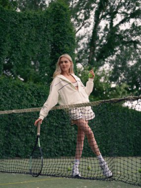 Elinde raket ve tenis topuyla tenis kortunda arka plan ve yeşil ağaçlarla antrenman yapan genç bir kadın için alt manzara. Aktif eğlence ve sağlıklı yaşam tarzı.