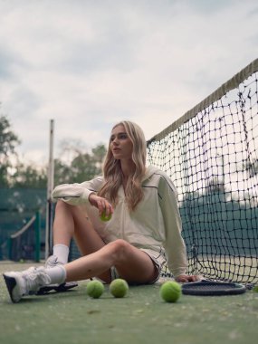 Genç atletik kadın tenis antrenmanından sonra dışarıda, elinde raket ve toplarla ağda oturuyor. Odaklanmış ve yorgun görünüyor..