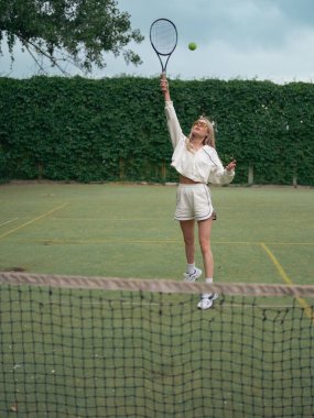 Spor giyimli, elinde raket, tenis kortunda tenis topu eğitimi alan güneş gözlüklü genç bir kadın tenis ağının arka planına karşı. Aktif eğlence ve sağlıklı yaşam tarzı.