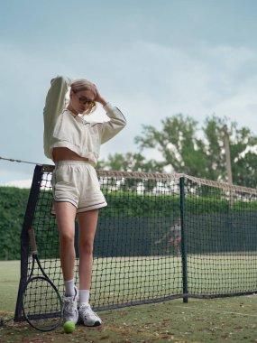 Spor giyim ve güneş gözlüklü genç bayan tenis antrenmanından sonra dışarıda raket ve topun yanında duruyor..