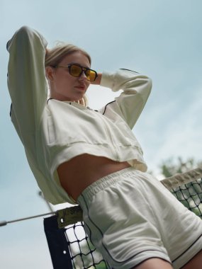 Spor giyim ve güneş gözlüklü genç atletik bayana alt manzara açık sahadaki tenis antrenmanından sonra dinleniyor, ağın yanında duruyor. Yakın plan..