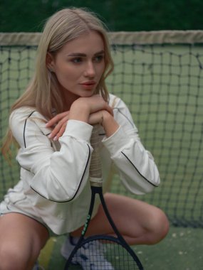 Spor giyimli genç bir kadının portresi. Tenis antrenmanından sonra dışarıda dinleniyor. Elinde raketle ağda oturuyor..