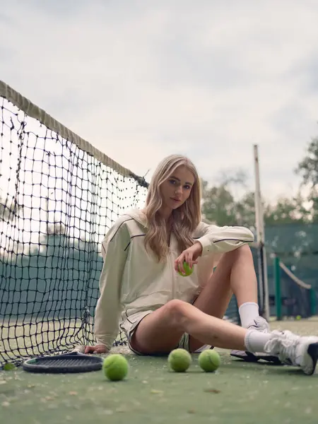 Genç atletik kadın tenis antrenmanından sonra dışarıda, elinde raket ve toplarla ağda oturuyor. Odaklanmış ve yorgun görünüyor..