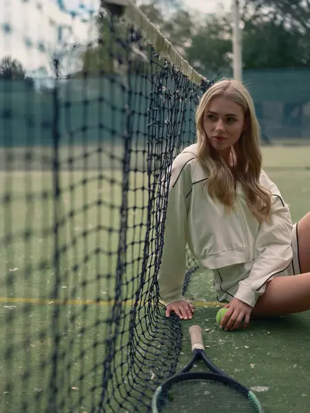 Genç atletik kadın tenis antrenmanından sonra dışarıda, elinde raket ve toplarla ağda oturuyor. Odaklanmış ve yorgun görünüyor..