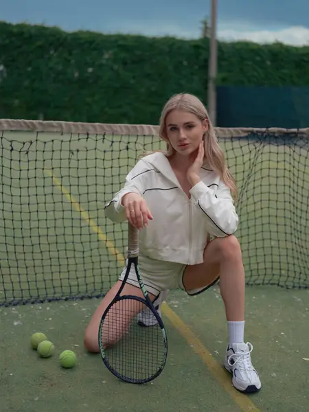 Genç atletik kadın tenis antrenmanından sonra dışarıda, elinde raket ve toplarla ağda oturuyor. Kameraya bakıyor..