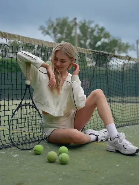 Genç gülümseyen atletik kadın tenis antrenmanından sonra elinde raketle ağ ve toplarla dışarıda dinleniyor..