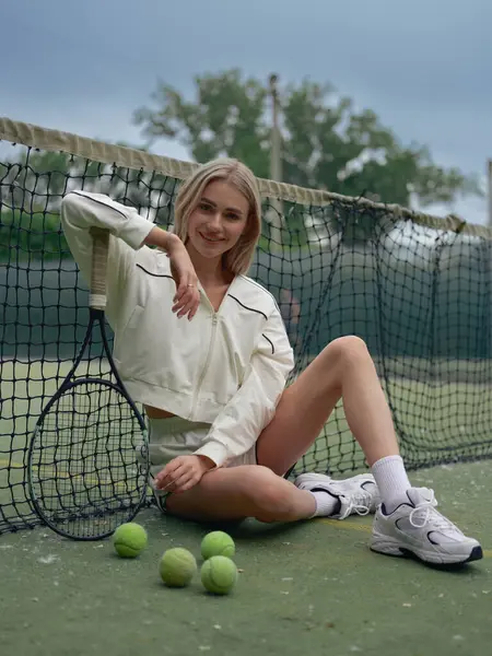 Genç gülümseyen atletik kadın tenis antrenmanından sonra elinde raketle ağ ve toplarla dışarıda dinleniyor..