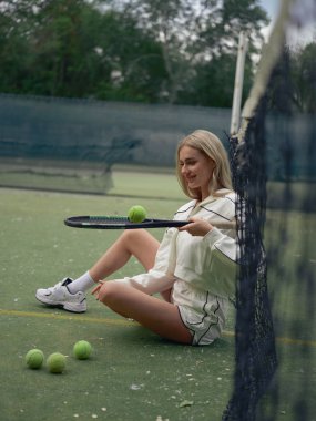 Genç gülümseyen atletik kadın tenis antrenmanından sonra dinleniyor. Ağ başında oturuyor ve raket topa vuruyor..