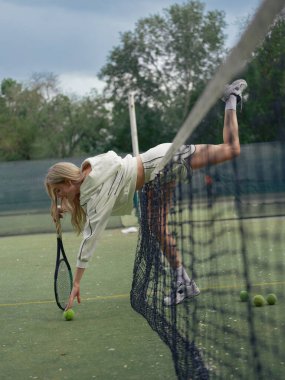 Elinde raketi olan genç bir kadın topa uzanıyor ve top için eğiliyor ve tenis kortunda becerilerini geliştiriyor. Aktif eğlence ve sağlıklı yaşam tarzı.