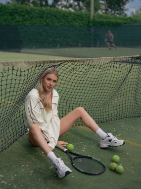 Genç atletik kadın tenis antrenmanından sonra dışarıda, elinde raket ve toplarla ağda oturuyor. Yorgun görünüyor. Üst görünüm.
