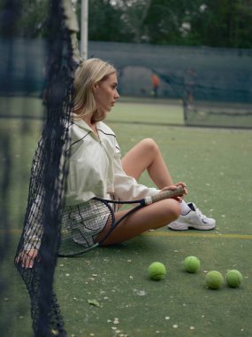 Genç atletik kadın tenis antrenmanından sonra dışarıda, elinde raket ve toplarla ağda oturuyor. Yan görünüm.