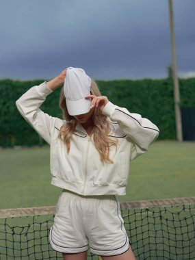 Genç sarışın bir kadın tenis kortunda spor kıyafetleri içinde duruyor ve beyaz şapka takıyor. Tenis eğitimi, aktif eğlence ve sağlıklı yaşam tarzı. Yakın plan..