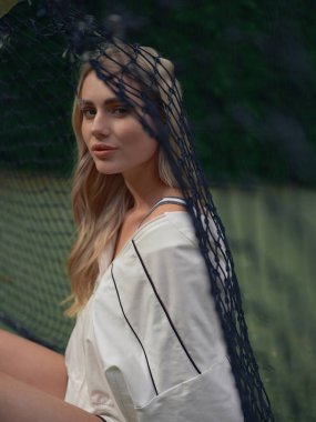 Spor kıyafetli genç bir kadının portresi. Tenis antrenmanından sonra dışarıda, ağın kenarında oturuyor..