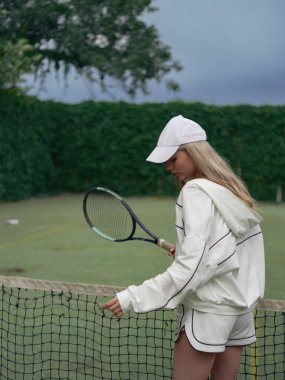 Elinde raketi olan genç bir atlet kadın tenis kortunda ağ, yeşil ağaçlar ve gökyüzü ile antrenman yapıyor. Tenis eğitimi, aktif eğlence ve sağlıklı yaşam tarzı. Yakın plan..