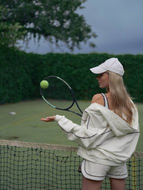 Elinde raketle tenis kortunda antrenman yapan genç bir atlet kadın. Arka planı ağ, yeşil ağaçlar ve gökyüzü ile çevrili. Tenis eğitimi, aktif eğlence ve sağlıklı yaşam tarzı. Yakın plan..