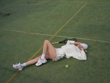 Genç yorgun atletik kadın tenis antrenmanından sonra dışarıda dinleniyor ve raket ve topların yanındaki spor sahasına uzanıyor. Tenis antrenmanı, aktif eğlence ve sağlıklı yaşam tarzı. Üst görünüm.