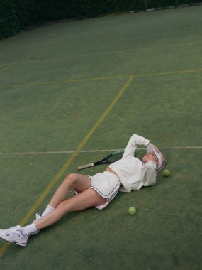 Genç yorgun atletik kadın tenis antrenmanından sonra dışarıda dinleniyor ve raket ve topların yanındaki spor sahasına uzanıyor. Tenis antrenmanı, aktif eğlence ve sağlıklı yaşam tarzı. Üst görünüm.