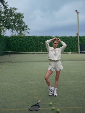 Spor giyimli genç bir bayan tenis antrenmanından sonra dışarıda dinleniyor ve raket ve topların arka planında poz veriyor..