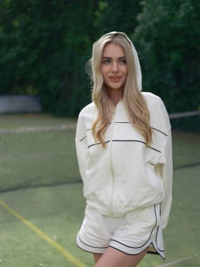 Kapüşonlu ceketli genç atletik kadın tenis antrenmanından sonra spor sahasının arka planında dinleniyor. Yakın plan..