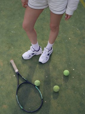 Spor ayakkabılı, raket ve tenis toplarının yanında tenis kortunda antrenman yapan atlet bir kadının bacaklarına yakın çekim. Aktif yaşam tarzı, açık hava sporu, tenis kortu ve tenis eğitimi.