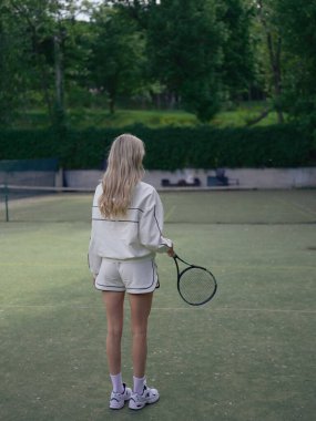 Elinde raketle tenis kortunda yeşil ağaçlarla antrenman yapan genç bir atlet kadın. Tenis eğitimi, aktif eğlence ve sağlıklı yaşam tarzı. Arka plan.