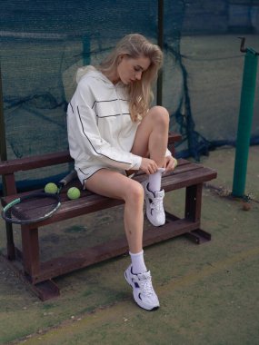 Genç atletik kadın antrenmandan önce tenis kortunda raket ve topların yanında bankta oturuyor, beyaz çoraplarını ayarlıyor ve yukarı çekiyor. Aktif yaşam tarzı, açık hava sporu, tenis eğitimi.