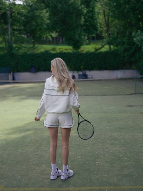 Elinde raketle tenis kortunda yeşil ağaçlarla antrenman yapan genç bir atlet kadın. Tenis eğitimi, aktif eğlence ve sağlıklı yaşam tarzı. Arka plan.