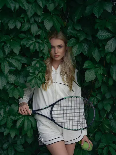 Elinde tenis raketi ve topları olan genç atlet kadın yemyeşil yapraklar ve genç üzüm filizleri arasında duruyor. Aktif eğlence ve sağlıklı yaşam tarzı. Yakın plan..