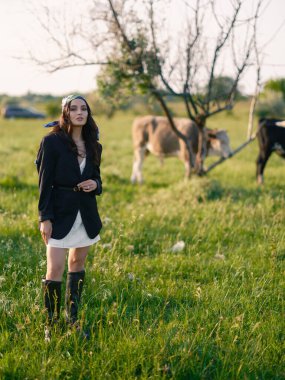 Kırsalda yeşil bir çayırda inekleri güden genç bir kadın. Çiftlik hayvanları, doğa ve tarım hayatıyla güzel manzaraya karşı kırsal yaz manzarası.