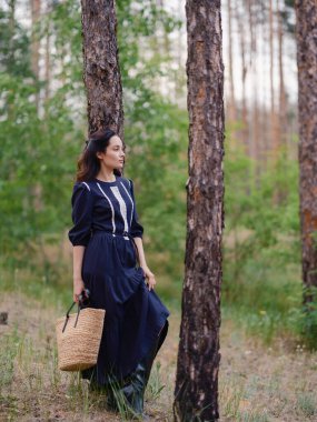 Siyah mor elbiseli, elinde saman torbası olan ve ormanda çam ağacı gövdeleri arasında duran düşünceli genç bir kadın. Çam ağaçlarıyla sakin doğal ortam, huzurlu atmosfer ve yumuşak gün ışığı..