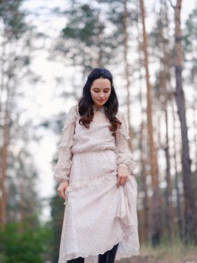 Açık renk elbiseli genç bir kadın çam ağaçlarının arasında dikiliyor. Yakın plan. Çam ağaçlarıyla sakin doğal ortam, huzurlu atmosfer ve yumuşak gün ışığı..