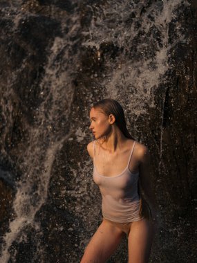 Beyaz bluzlu ve bikinili genç ıslak kadın kayaların arasında, şelalenin altında duruyor. Huzurlu doğal ortam ve yumuşak ışık. Yaz günü, doğa bağlantısı ve açık hava rahatlaması..