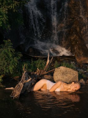 Genç, ıslak kadın, sığ bir ormanın kenarında şelaleye karşı ağaç gövdesi ve taşın yanında uzanıp dinleniyor. Doğal ortam, yumuşak ışık. Yaz günü, doğa bağlantısı ve açık hava rahatlaması..