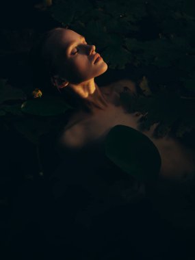 Gölde yüzen, yaprakların ve sarı nilüferlerin arasında dinlenen genç bir kadının portresi. Yakın plan. Üst Manzara. Yaz havası, rahatlama ve doğal güzellik..