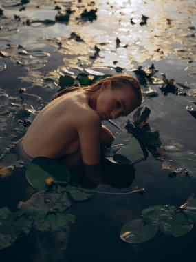 Genç bir kadın gölde oturuyor ve yaprakların ve sarı nilüferlerin arasında suda dinleniyor. Arka ışıklandırma. Yakın plan. Yaz havası, rahatlama ve doğal güzellik..