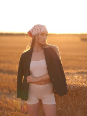 Bandana ve siyah ceket giyen, gün batımında altın hasat edilmiş buğday tarlasında saman balyasının yanında poz veren beyaz etekli ve şortlu şık bir kadın. Sıcak kırsal yaz atmosferi. Yakın plan. Arka ışıklandırma.
