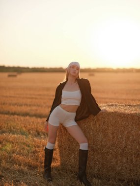 Bandana ve siyah ceket giyen, gün batımında altın hasat edilmiş buğday tarlasında saman balyasının yanında poz veren beyaz etekli ve şortlu şık bir kadın. Sıcak kırsal yaz atmosferi. Arka ışıklandırma.