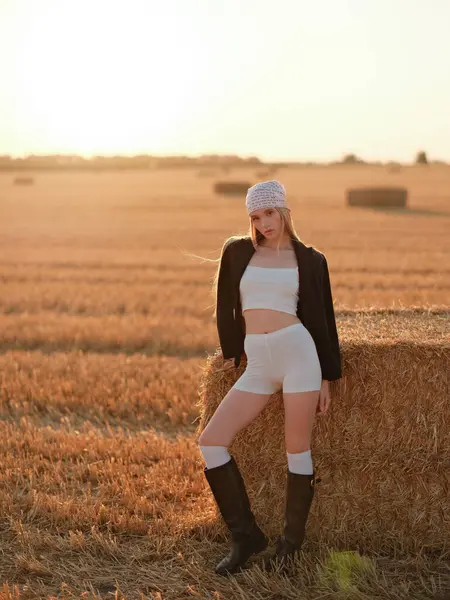 Bandana ve siyah ceket giyen, gün batımında altın hasat edilmiş buğday tarlasında saman balyasının yanında poz veren beyaz etekli ve şortlu şık bir kadın. Sıcak kırsal yaz atmosferi.