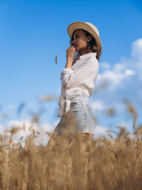 Hasır şapkalı, beyaz gömlekli ve kot şortlu genç bir kadın mavi gökyüzünün altındaki altın buğday tarlasında duruyor. Doğal güzellik ve güneş ışığıyla huzurlu kırsal bir yaz manzarası. Alt görünüm.