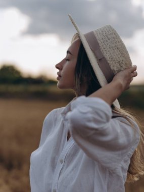 Hasır şapkalı ve beyaz gömlekli genç kadın buğday tarlasında gözleri kapalı dinleniyor ve dinleniyor. Huzurlu kırsal yaz manzarası, doğal güzellik. Yakın plan. Portre.
