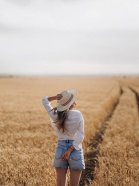 Hasır şapkalı, beyaz tişörtlü ve kot şortlu genç bir kadın buğday tarlasına giden yolda duruyor. Doğal güzellik ve güneş ışığıyla huzurlu kırsal bir yaz manzarası. Arka plan.