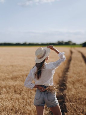 Hasır şapkalı, beyaz tişörtlü ve şortlu genç bir kadın buğday tarlasına doğru giden yolda buğday kontu sürüsüyle duruyor. Huzurlu kırsal yaz manzarası, doğal güzellik. Arka plan.